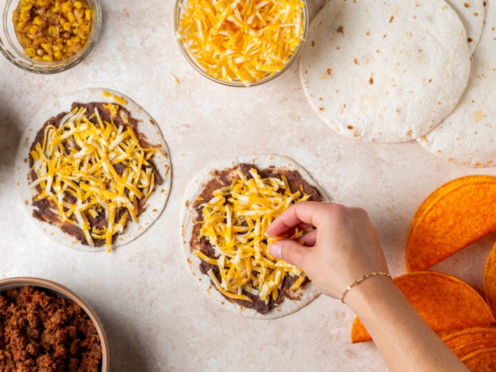 Shredded colby jack cheese being sprinkled over refried beans on two flour tortillas.