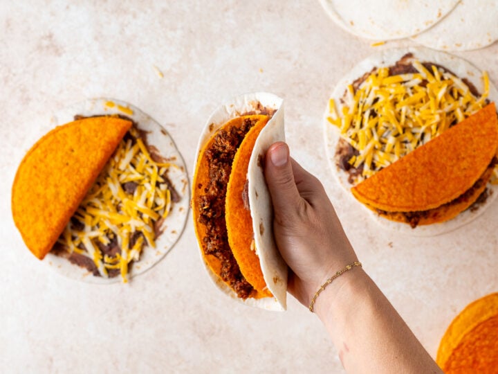 Hand holding an assembled crunchy taco wrapped in a soft flour tortilla with refried beans and cheese.