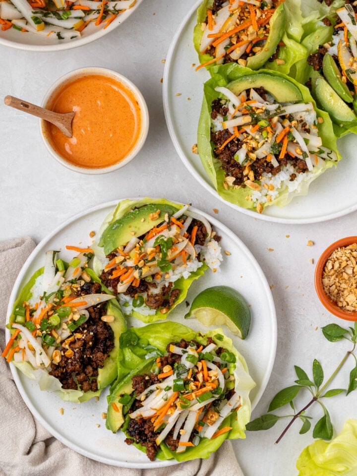 Two white plates full of lettuce leaves with rice, gochujang ground beef, pickled pear slaw, avocado, and peanuts