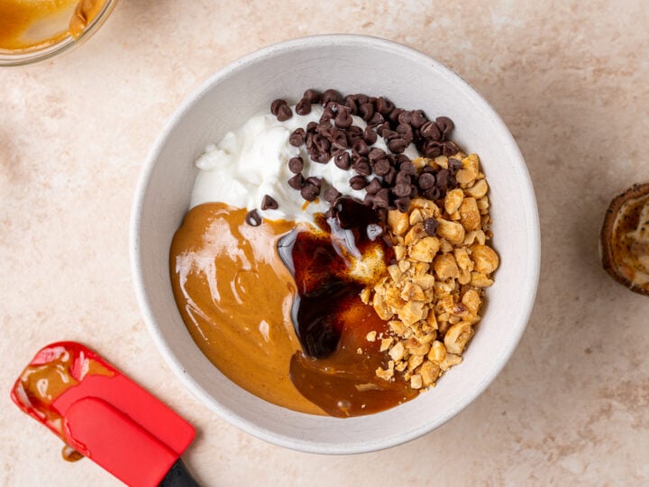 Greek yogurt, peanut butter, salted caramel syrup, vanilla extract, chocolate chips and chopped peanuts added to a bowl.
