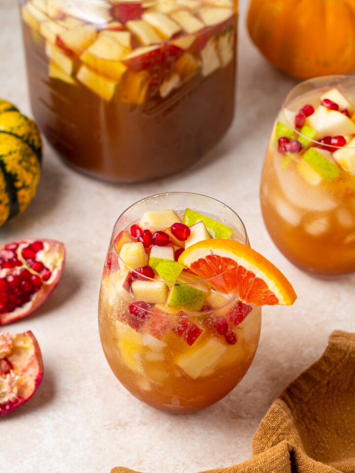 Close-up of a glass of sparkling apple cider sangria garnished with an orange slice, with another glass and pitcher in the background.