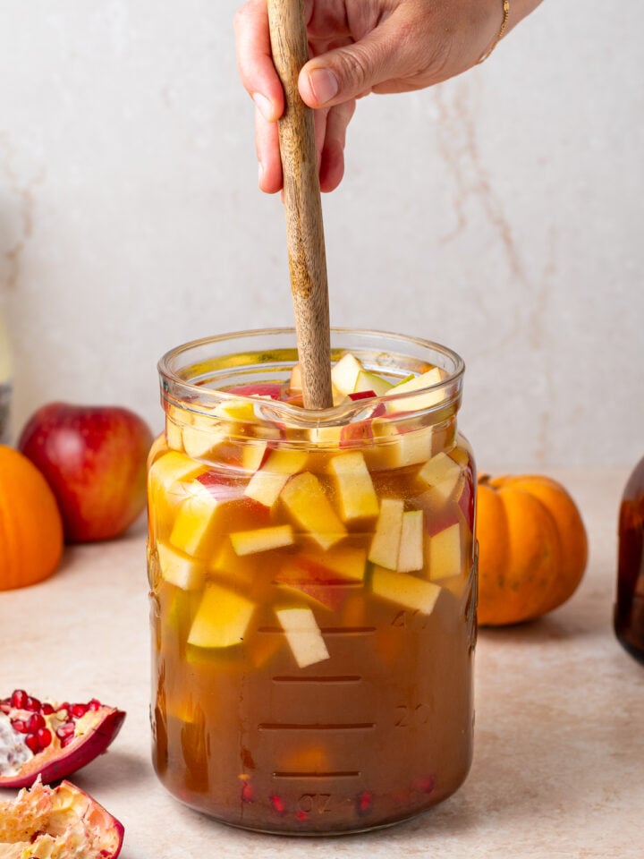 Stirring a large pitcher of fall white sangria with a wooden spoon to combine the fruit and liquids.