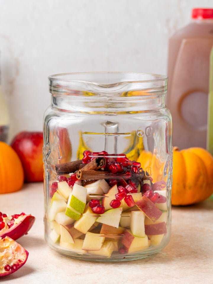 Chopped apples, pears, pomegranate arils and cinnamon sticks in a large glass pitcher.