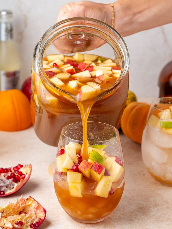 Pouring apple cider sangria from a large glass pitcher into a stemless wine glass filled with ice and fresh fruit.