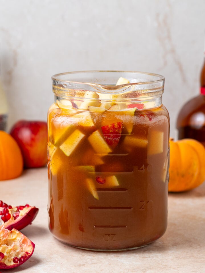 Glass pitcher filled with apple cider sangria, chopped apples, pears, pomegranate arils and cinnamon sticks.