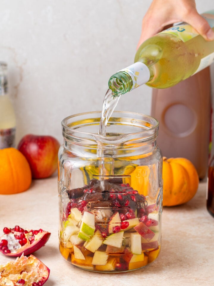 Pouring white wine into a pitcher with chopped apples, pears, pomegranate seeds and cinnamon.