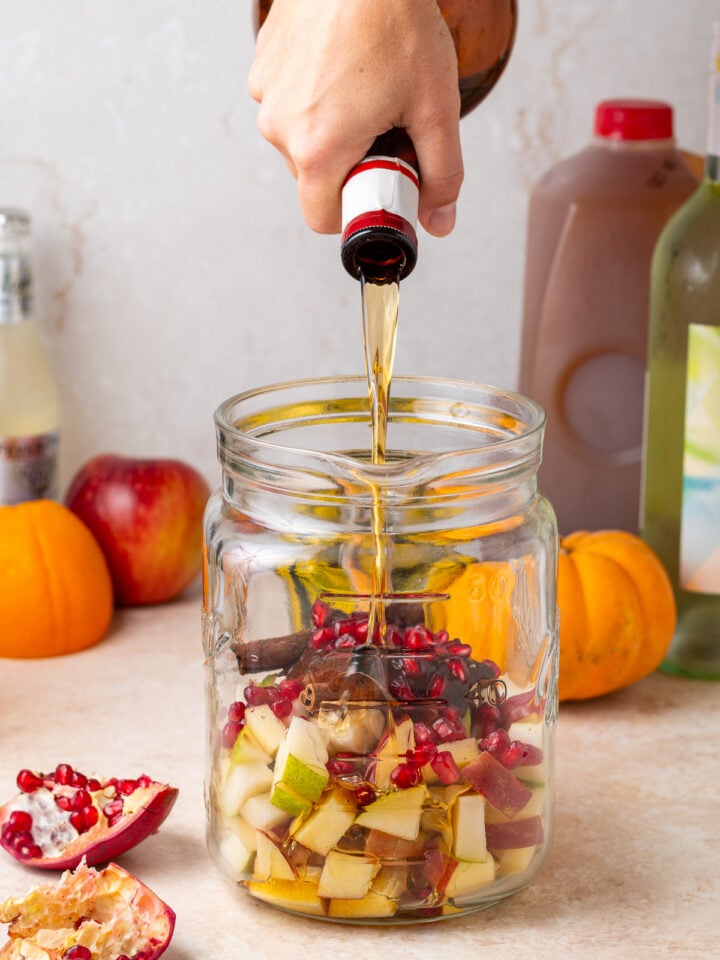 Pouring Grand Marnier into a glass pitcher filled with chopped fall fruit and cinnamon sticks.